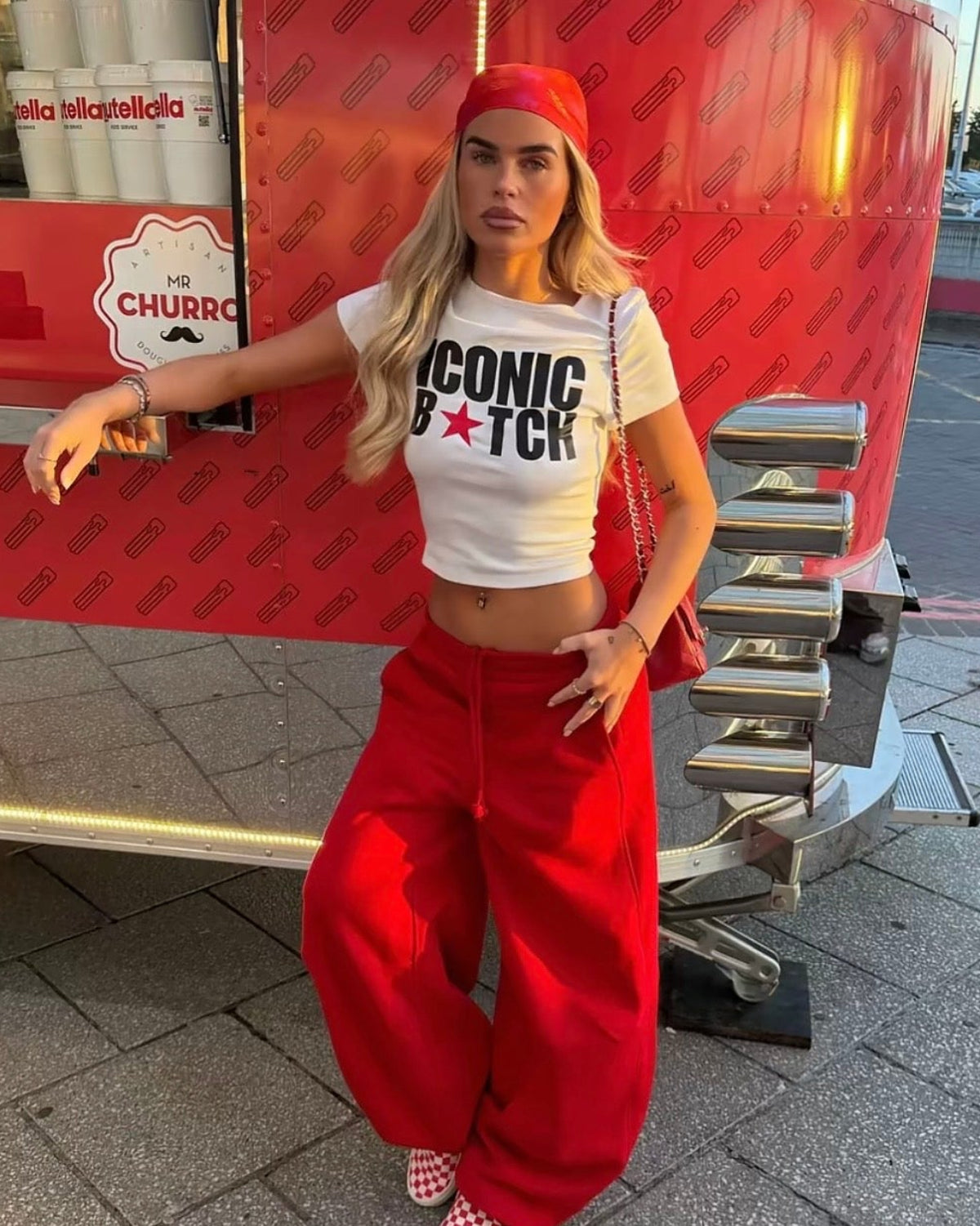 A woman posing in front of a red food truck, wearing a white graphic crop top that reads 'ICONIC B*TCH' with a red star, red wide-leg sweatpants, a red beanie, and red-and-white checkered sneakers.