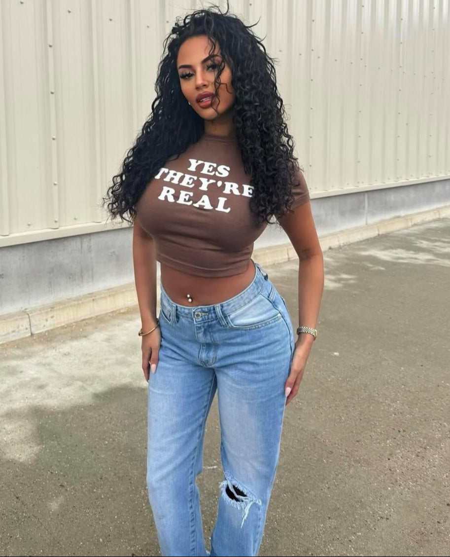 Woman wearing a brown crop top with text and light blue jeans standing against a white wall.